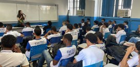 Sala de aula com estudantes uniformizados sentados e atentos, enquanto a palestrante fala ao mic...