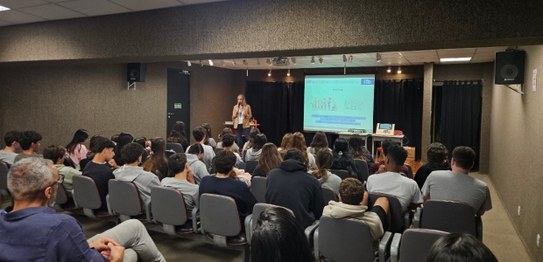 Auditório com cortina vermelha ao fundo, onde um homem está à frente, conduzindo uma apresentaçã...