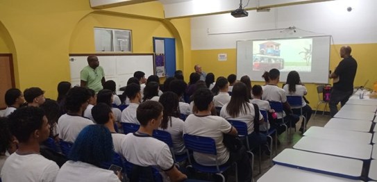 Um palestrante fala para jovens sentados em cadeiras azuis em um auditório simples, com bandeira...
