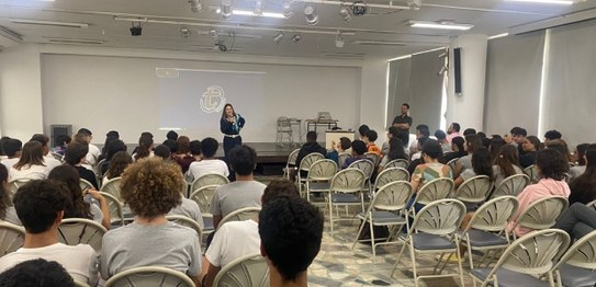 Auditório com cortina vermelha ao fundo, onde um homem está à frente, conduzindo uma apresentaçã...