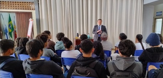 Um palestrante fala para jovens sentados em cadeiras azuis em um auditório simples, com bandeira...