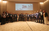 Foto posada das(os) participantes, que estão de pé, formando um semi-círculo; tendo ao funo a lo...