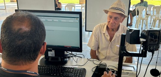 Foto do atendimento de um homem idoso, ele usa chapéu de palha e camisa clara. A câmera está foc...