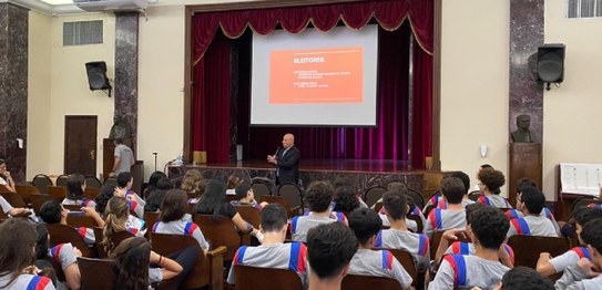 Numa sala de aula, sentados, estudantes assistem à palestra do juiz eleitoral. Vestindo terno pr...