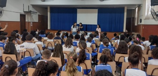 Numa sala de aula, sentados, estudantes assistem à palestra do juiz eleitoral. Vestindo terno pr...