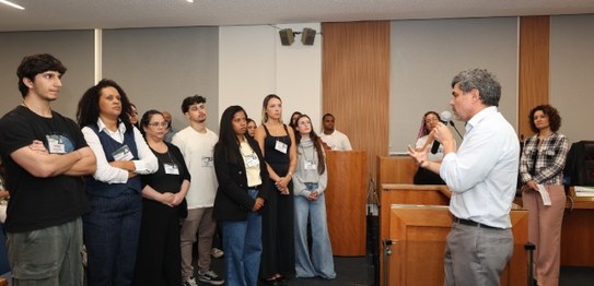 Um homem está de pé em um auditório, segurando um microfone e falando para um grupo de alunos se...