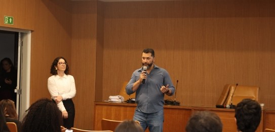 Um homem está de pé em um auditório, segurando um microfone e falando para um grupo de alunos se...