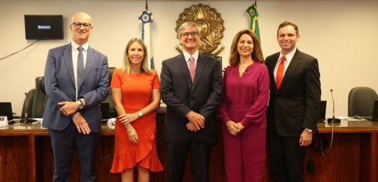 Foto posada de participantes do evento, tendo ao fundo o brasão da República e bandeira do Brasi...