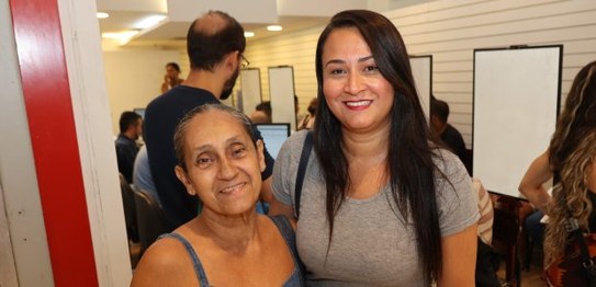 Mulher parda, de óculos e cabelos curtos, posa sorrindo e fazendo sinal de positivo; ao fundo, g...