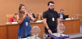 Uma mulher segura um microfone e gesticula com a mão enquanto fala ao lado de um homem de camisa...
