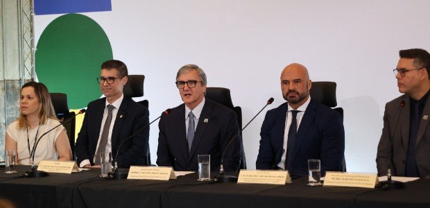 Descrição da imagem: Na coletiva de imprensa, da esquerda para a direita, estão sentados, a dire...