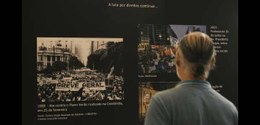 Um homem, de costas, está olhando um dos painéis da exposição que contém fotos históricas do cen...