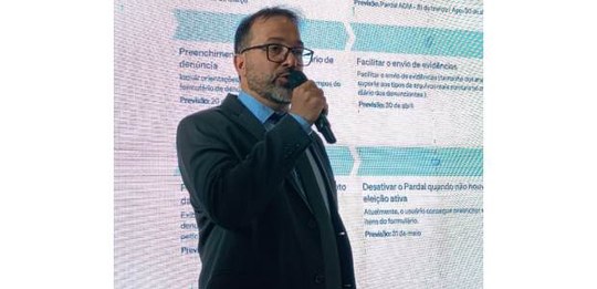 Foto de Pablo de Barros falando ao microfone; ao fundo projeção com textos; ele usa óculos e ves...