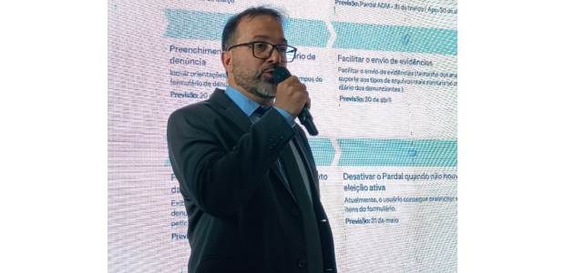 Foto de Pablo de Barros falando ao microfone; ao fundo projeção com textos; ele usa óculos e ves...