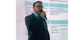 Foto de Pablo de Barros falando ao microfone; ao fundo projeção com textos; ele usa óculos e ves...