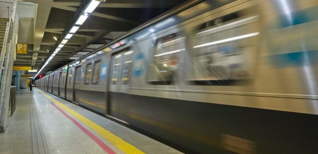 A imagem mostra um trem de metrô em movimento numa plataforma subterrânea. O trem está em alta v...