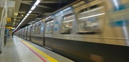 A imagem mostra um trem de metrô em movimento numa plataforma subterrânea. O trem está em alta v...