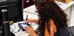 Mulher sentada diante de um computador utilizando uma mesa digitalizadora/caneta eletrônica para...