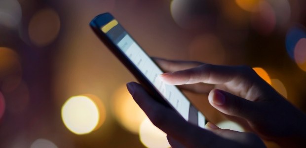 TRE-RJ Celular mão manipula a tela de um celular; ao fundo, pontos de luz desfocados