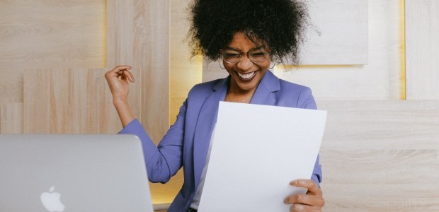 Foto de uma mulher negra segurando um papel diante de um notebook; ela veste terno lilás e usa ó...