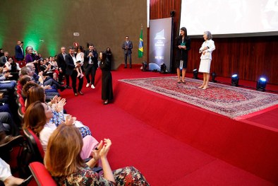 A imagem mostra uma cerimônia em auditório, com plateia aplaudindo enquanto um grupo de pessoas ...