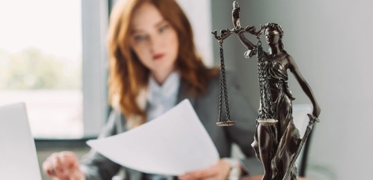 Mulher sentada em um escritório analisa documentos enquanto, em primeiro plano, aparece uma está...