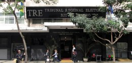 Descrição da imagem: Panorama da fachada da sede do TRE-RJ, um prédio de mármore escuro, com pes...