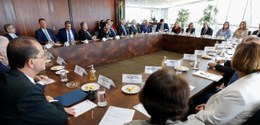 A imagem retrata uma reunião com várias pessoas sentadas ao redor de quatro mesas longas, organi...