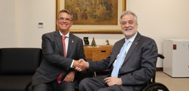 Dois homens de terno estão sentados em um ambiente de escritório e apertam as mãos, sorrindo par...