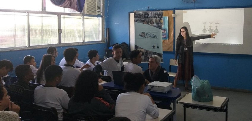 Em pé, na frente da sala de aula, a servidora Isabela Feijó aponta para o quadro branco, durante...