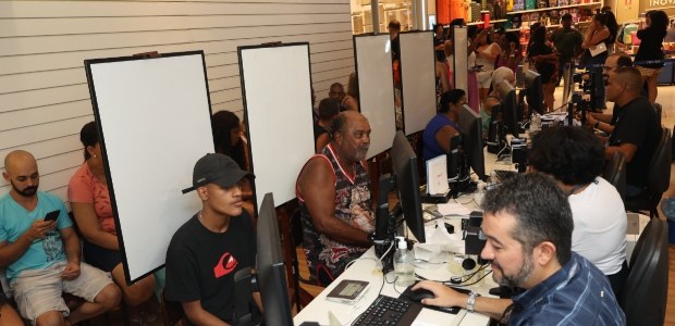 TRE-RJ faz atendimento itinerante com foco em biometria, no Bangu Shopping Guichês de atendimento, onde pessoas estão sentadas em frente a servidores, que estão sentados ...