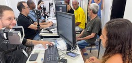 Balcão de atendimento, onde estão quatro pessoas sentadas à mesa. Do outro lado da mesa, onde há...