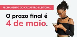 Mulher jovem e de cor da pele preta olha para o celular, ela usa óculos e uma blusa preta. Do la...