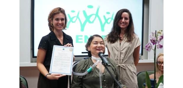 Três mulheres posam juntas em um evento; uma delas está sentada com um microfone e equipamento m...