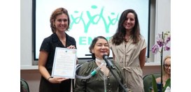 Três mulheres posam juntas em um evento; uma delas está sentada com um microfone e equipamento m...