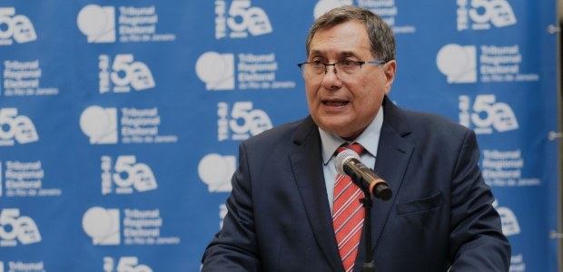Desembargador Peterson Barroso Simão discursa, tendo ao fundo um backdrop azul, com logo da Just...