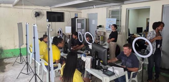 Ambiente com quatro guichês de atendimento, com mesas de plástico; as pessoas, vestindo blusa am...