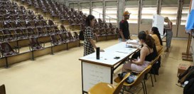A foto mostra um auditório amplo com arquibancadas repletas de cadeiras marrons e algumas amarel...