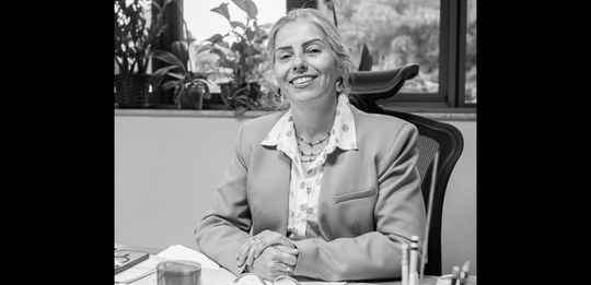Imagem em preto e branco, que mostra uma mulher sorrindo, sentada atrás de uma mesa em um ambien...