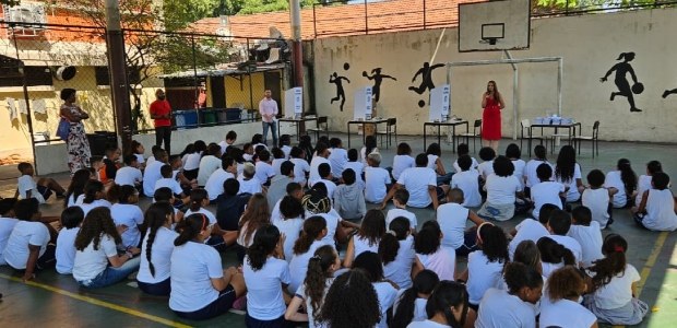 TRE-RJ Programa “TRE vai à Escola” visitou quatro escolas da Capital em setembro Descrição da imagem: Na quadra da escola, a juíza está em pé, segura um microfone e veste um ves...