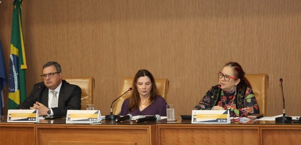 A imagem mostra a mesa de autoridades do evento, realizado no auditório. Três pessoas estão sent...