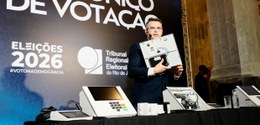 O secretário de TI apresenta uma urna eletrônica aberta, exibindo seus componentes internos, sob...