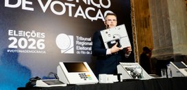 O secretário de TI apresenta uma urna eletrônica aberta, exibindo seus componentes internos, sob...