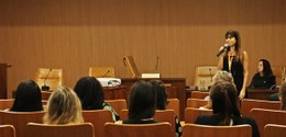 Em um auditoria, pessoas estão sentadas e assistem a servidora de roupa preta e com o microfone ...