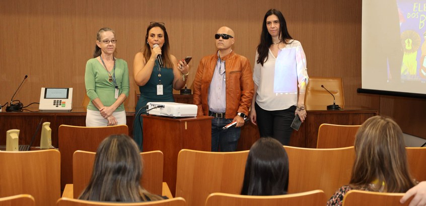 TRE-RJ recebe estudantes do Instituto Sorrindo para ação educativa sobre o processo eleitoral A imagem mostra um grupo de quatro pessoas em pé, de frente para uma plateia. Uma das pessoas es...