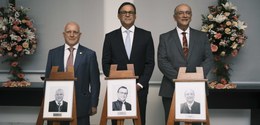 A imagem mostra três homens de terno e gravata posando para uma foto em um ambiente formal, poss...