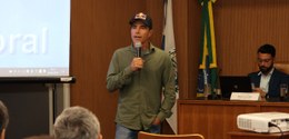 O surfista está em pé, falando ao microfone. Ele veste uma camisa verde de manga comprida, calça...