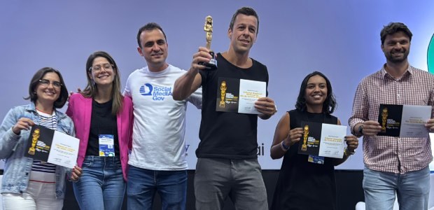 TRE-RJ vence prêmio nacional de comunicação pública A imagem mostra um grupo de seis pessoas em um palco durante uma cerimônia de premiação. Todos e...