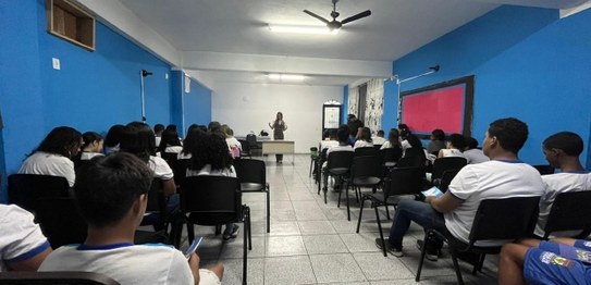 Auditório com cortina vermelha ao fundo, onde um homem está à frente, conduzindo uma apresentaçã...