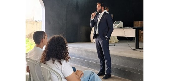 Alunos sentados assistem palestra feita por homem de terno, que aponta para duas urnas eletrônic...